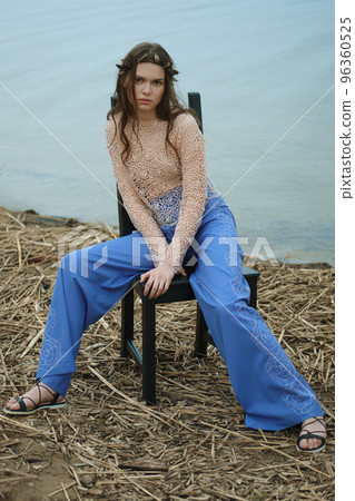 Full length portrait of young woman in blue pants and lace blouse sitting on chair shore of the lake Full length portrait of young woman in blue pants and lace blouse sitting on chair shore of the lake 96360525