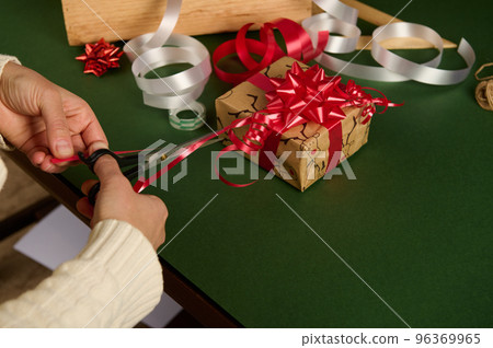 Close-up of a woman cutting shiny ribbon in strips for making beautiful ornaments while wrapping presents for Christmas Close-up of a woman cutting shiny ribbon in strips for making beautiful ornaments while wrapping presents for Christmas 96369965