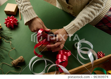 Top view of a woman wrapping Christmas gift, tying a bow from a red shiny decorative ribbon. Boxing Day. Diy presents 96369974