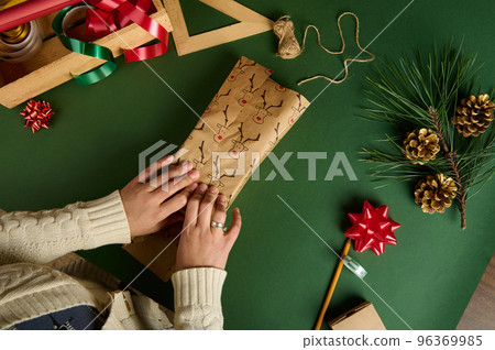 Top view of a woman packing present in decorative paper with deer pattern, over green background with wrapping materials 96369985