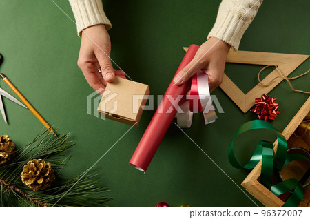 Woman holds small cardboard box, a red wrapping paper and decorative shiny ribbons for packing present at green surface Woman holds small cardboard box, a red wrapping paper and decorative shiny ribbons for packing present at green surface 96372007