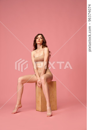 Portrait of young beautiful woman with slim muscular body sitting, posing in beige underwear isolated over pink studio background Portrait of young beautiful woman with slim muscular body sitting, posing in beige underwear isolated over pink studio background 96374074