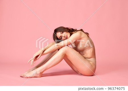 Portrait of young beautiful woman posing, sitting on floor in beige underwear isolated over pink studio background. Slender body, smooth legs Portrait of young beautiful woman posing, sitting on floor in beige underwear isolated over pink studio background. Slender body, smooth legs 96374075