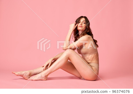 Portrait of young beautiful woman posing in beige underwear, sitting on floor isolated over pink studio background. Smooth legs. Depilation and epilation Portrait of young beautiful woman posing in beige underwear, sitting on floor isolated over pink studio background. Smooth legs. Depilation and epilation 96374076