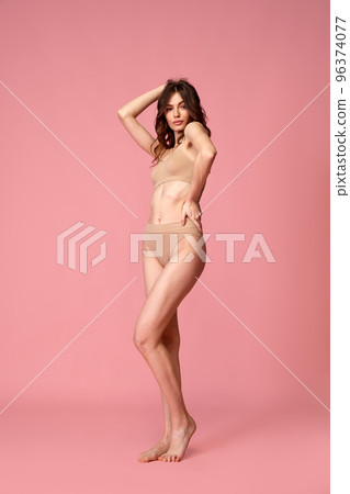 Portrait of young beautiful woman with brunette curly hair posing in beige underwear isolated over pink studio background. Slender body Portrait of young beautiful woman with brunette curly hair posing in beige underwear isolated over pink studio background. Slender body 96374077