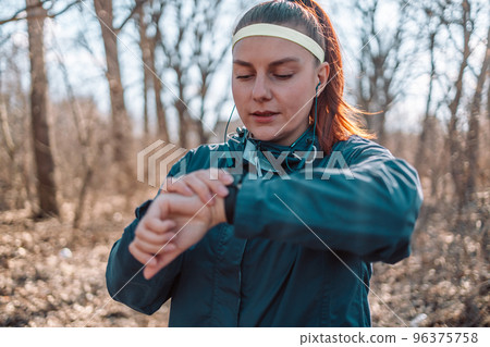 Sport watch run woman checking smartwatch tracker. Trail running runner girl looking at heart rate monitor smart watch in forest wearing jacket sportswear. Female athlete jogger training in woods. Sport watch run woman checking smartwatch tracker. Trail running runner girl looking at heart rate monitor smart watch in forest wearing jacket sportswear. Female athlete jogger training in woods. 96375758