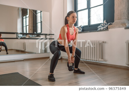 A strong, muscular female weightlifter is doing squats with a kettlebell in a gym. A strong, muscular female weightlifter is doing squats with a kettlebell in a gym. 96375849