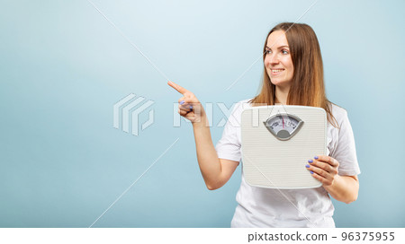 Woman with weighing scale pointing with finger to side on blue background 96375955