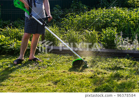 Defocus grass trimmer. A man mowing the grass. Outdoor view of young worker using a lawn trimmer mower cutting grass in a blurred nature background. Housework. Out of focus 96378584