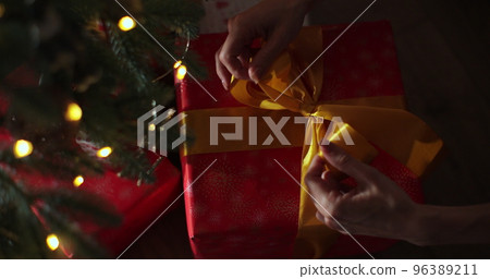 Hands of woman using ribbon to tie bow for a wrapped gift box for Christmas present with Xmas tree in foreground and colorful lights bokeh. Hands of woman using ribbon to tie bow for a wrapped gift box for Christmas present with Xmas tree in foreground and colorful lights bokeh. 96389211