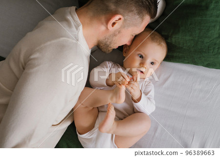 Cheerful, beautiful newly-made dad and a charming six-month-old baby lie together on green and white underwear. A young father with a small child in a home portrait. Top view 96389663