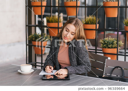 Young charming woman is calling on a mobile phone, sitting in a cafe and talking on a mobile phone with a smile, writing notes in a notebook. Remote work Young charming woman is calling on a mobile phone, sitting in a cafe and talking on a mobile phone with a smile, writing notes in a notebook. Remote work 96389664