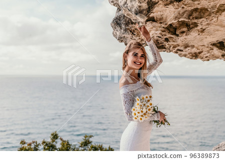 Romantic bride, blond girl in white wedding dress with open shoulders posing with sea and rocks in background. Stylish young woman standing on cliff's edge and looking into a ocean mountains at sunset Romantic bride, blond girl in white wedding dress with open shoulders posing with sea and rocks in background. Stylish young woman standing on cliff's edge and looking into a ocean mountains at sunset 96389873
