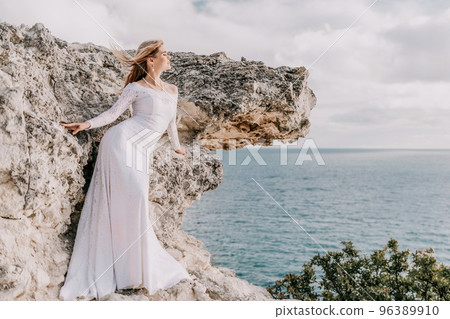 Romantic bride, blond girl in white wedding dress with open shoulders posing with sea and rocks in background. Stylish young woman standing on cliff's edge and looking into a ocean mountains at sunset Romantic bride, blond girl in white wedding dress with open shoulders posing with sea and rocks in background. Stylish young woman standing on cliff's edge and looking into a ocean mountains at sunset 96389910