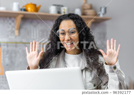 Young beautiful Latin American woman talking on video call at home in the kitchen. She is happy talking, saying hello, waving her hands. Young beautiful Latin American woman talking on video call at home in the kitchen. She is happy talking, saying hello, waving her hands. 96392177