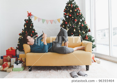 A single man seems lonely using his phone and lay down on sofa in living room decorated with Christmas trees and gift boxes 96393464