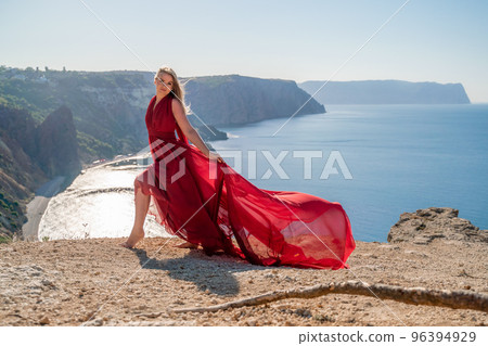 A woman in a red flying dress fluttering in the wind, against the backdrop of the sea. A woman in a red flying dress fluttering in the wind, against the backdrop of the sea. 96394929