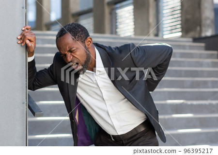 African american man overworked outside office building, boss in business suit has severe back pain, businessman near stairs holding hands on back muscles. 96395217