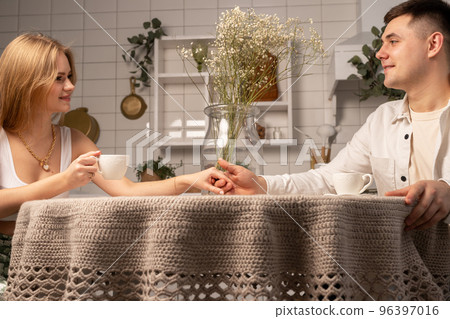 Happy couple sitting at kitchen table, drinking tea or coffee, holding hands, looking at each other. Cozy romantic atmosphere. High quality photo 96397016