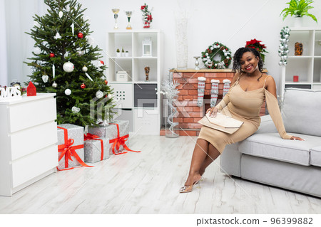 An African woman sits on a sofa in a beige dress in a room decorated for Christmas. An African woman sits on a sofa in a beige dress in a room decorated for Christmas. 96399882