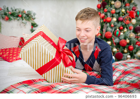 A cheerful teenager boy opens a Christmas gift. Cheerful Teenager lies in bed with a christmas present in his hands 96406693
