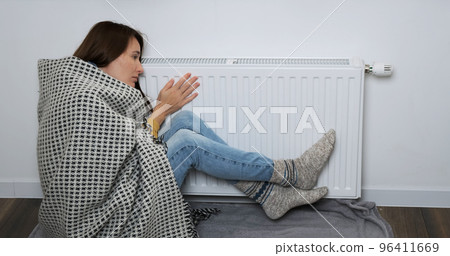 Young woman sitting near battery, warming herself. Woman wearing in warm socks, woolen blanket. Low temperature in apartment, house, room in cold autumn, winter concept. Poor central heating indoors 96411669