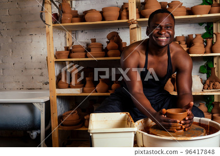 handsome and confident afro american man potter bare body apron naked sculpts clay vase . sexy sculptor african student In workshop on the background of a rack with pottery dishes. art do it yourself handsome and confident afro american man potter bare body apron naked sculpts clay vase . sexy sculptor african student In workshop on the background of a rack with pottery dishes. art do it yourself 96417848