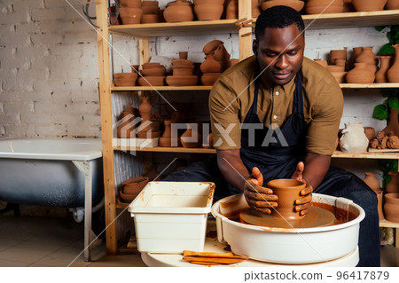 mixed race afro male potter with black apron and stylish dark shirt sitting at workshop table potter's wheel , applying glaze on handmade clay brown pot vase mixed race afro male potter with black apron and stylish dark shirt sitting at workshop table potter's wheel , applying glaze on handmade clay brown pot vase 96417879