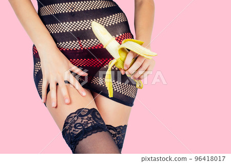 Closeup of a sexy woman body in red panties and black sexy transparent dress stockings touching a banana in a condom on pink background in the studio. intimate massage games cock safe sex 96418017