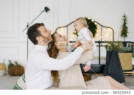 Young parents lift their baby in their arms and smile at him against the background of a bedroom decorated for Christmas and New Year Young parents lift their baby in their arms and smile at him against the background of a bedroom decorated for Christmas and New Year 96420843