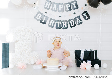 A one year old girl sits on the floor near a sweet cake in honor of her first birthday, tries the cake and smiles, laughs A one year old girl sits on the floor near a sweet cake in honor of her first birthday, tries the cake and smiles, laughs 96420845