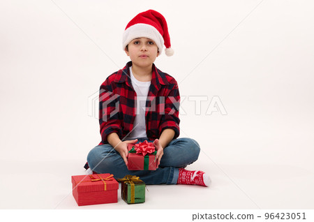 Full length portrait: handsome preteen boy in red plaid shirt and Santa hat, holding a Christmas gift, sitting on white background with copy space for text. Looking forward to upcoming winter holidays 96423051