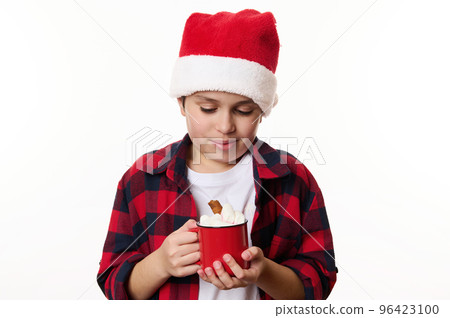 Adorable preteen boy wearing Santa hat, warming his hands with a red cup of hot chocolat and marshmallows, isolated on white background with copy advertising space. Merry Christmas, New Year concept Adorable preteen boy wearing Santa hat, warming his hands with a red cup of hot chocolat and marshmallows, isolated on white background with copy advertising space. Merry Christmas, New Year concept 96423100