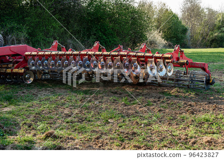 A red modern harrow stands in a field before sowing. Hydraulic mechanism for grinding the soil after plowing. A red modern harrow stands in a field before sowing. Hydraulic mechanism for grinding the soil after plowing. 96423827