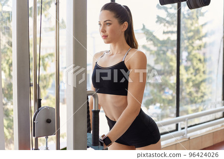 Athletic young woman doing exercises on simulator for upper muscle groups, gets herself in shape, develops strength. Attractive femalle with pony tail and black sport wear works out in gymnasium. 96424026