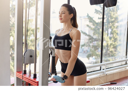 Portrait of magnetic sexy sporty young focused female doing strength exercises, being concentrated on result, wearing black sport top and shorts, having ponytail. Sport and motivation concept. 96424027