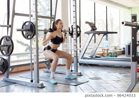 Image of professional sportswoman doing exercise in gym, squats with barbell, lifts weights, develops strength and definition of muscles, wears fasion sport uniform, has ponytail, keeps fit. 96424033