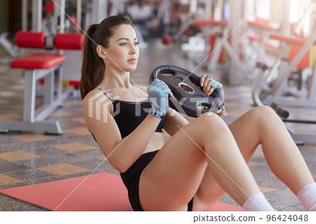 Close up of woman doing abs exercises with weight plate while sitting on sport mat on gym floor. Side view, sporty girl wears black stylish uniform, with ponitail. Healthy lifestyle concept. 96424048