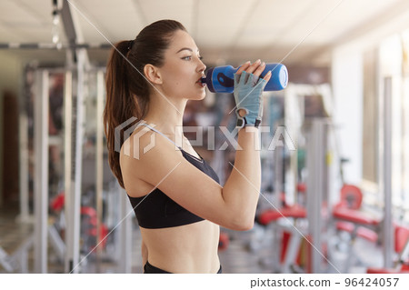 Half length shot of thirsty athletic woman drinking water at gym after working out, has slim perfect body shape, wears casual clothes and sportshoes, holds blue bottle. Sport and motivation concept. 96424057