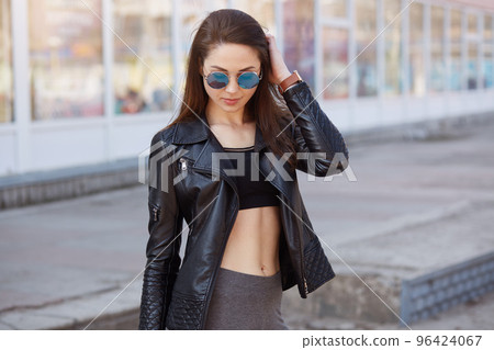 Image of attractive dark haired female wearing stylish leather jacket, bra and leggins, posing outdoors with bared belly, keeping hand on her beautiful hair, looking down. Lifestyle concept. 96424067