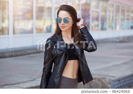 Closeup portrait of adorable confident female posing outdoors, wearing stylish clothing and sunglasses, looking in distance, standing with bared belly, keeps hand on her hair, expresses confidence. Closeup portrait of adorable confident female posing outdoors, wearing stylish clothing and sunglasses, looking in distance, standing with bared belly, keeps hand on her hair, expresses confidence. 96424068