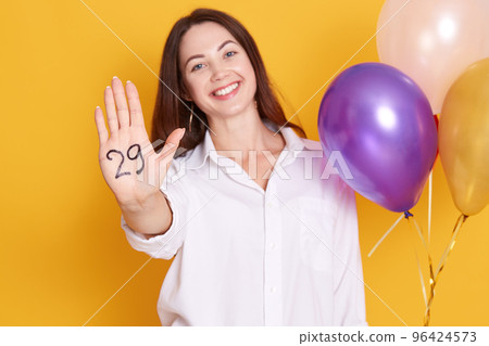 Portrait of happy smiling attractive dark haired woman with multi colored balloons in hand, showing her other palm with inscription twentie nine, female xpressing hapiness, celebrating with femily. 96424573