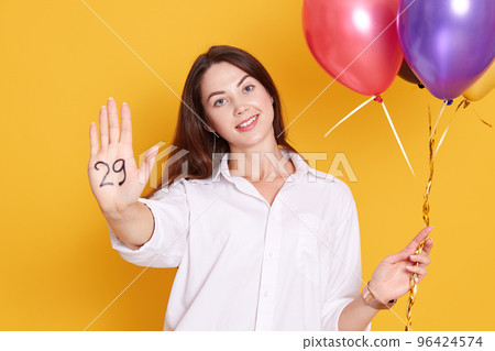 Studio shot of joyful young woman with party balloons in hand, celebrating her birthday, showing inscription twentie nine on her palm, wearing white elegant blouse, has dark hair, isolated on yellow. 96424574