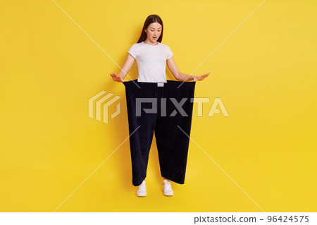 Full length photo of attractive young female shows her old huge pair of pants, looks shocked and surprised, looking down with opened mouth, lady after weight loss posing isolated on yellow background 96424575