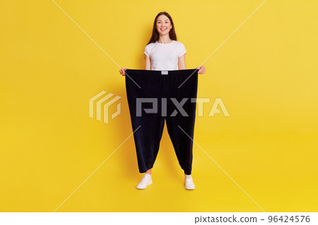 Young European female with dark hair wearing casual white t shirt and black pants too big size, adorable happy woman loose weight, feels great, expressing positive, keeps hands in trousers. 96424576