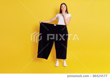 Full length of Caucasian astonished female lost weight and wearing old pants, being shocked and covering mouth with palm, posing isolated over yellow background. 96424577