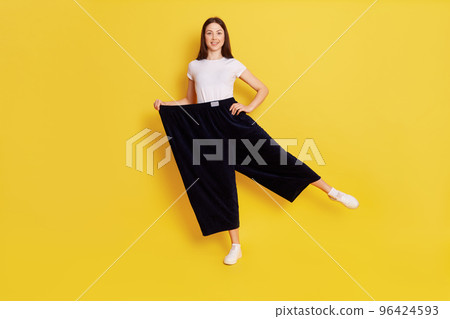 Happy woman after weight loss posing on one hand isolated over yellow background, wearing white casual t shirt and too big black pants, keeps one hand on hip, rejoices at achieved result. 96424593
