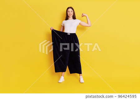 Full length shot of beautiful dark haired female wearing t shirt and old big size pants, showing loss weight results, raising hand to show muscles and her power, isolated over yellow background. 96424595
