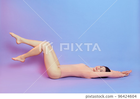 Full length photo of female with perfect body, lying on floor and stretching arms, raising long legs, dresses beige bodysuit, isolated over blue wall with pink neon light. 96424604