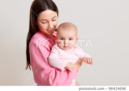 Dark haired Caucasian mother with long dark hair holding her cute baby girl wearing bodysuit, woman in casual outfit looking at her daughter with love, isolated over white background. 96424696